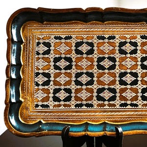 May include: Rectangular decorative tray with a scalloped edge. The tray features a gold border, a black and teal frame, and a patterned center with brown, black, and white geometric designs. The tray is displayed on a black stand.