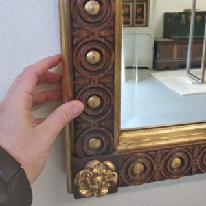 May include: A rectangular mirror with a gold frame and a dark brown, carved wooden border. The border has a repeating pattern of circles and gold accents. A hand is touching the frame.