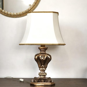 Puede incluir: Una lámpara de mesa con una base de metal en tono dorado con detalles tallados en volutas y una pantalla cuadrada de tela color blanco roto. La pantalla está rematada con un fino borde dorado en los bordes superior e inferior.
