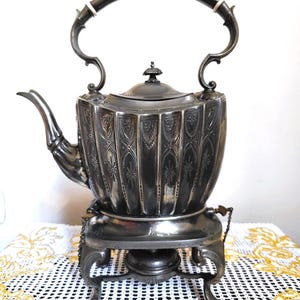 May include: A silver-toned antique tea kettle with a decorative floral pattern, a curved spout, and a large handle. The kettle sits on a matching stand with ornate legs and a small burner. The design is vintage and elegant.