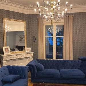 May include: A living room interior with a large gold chandelier, two blue velvet sofas, a glass coffee table, and a fireplace with a marble mantel.