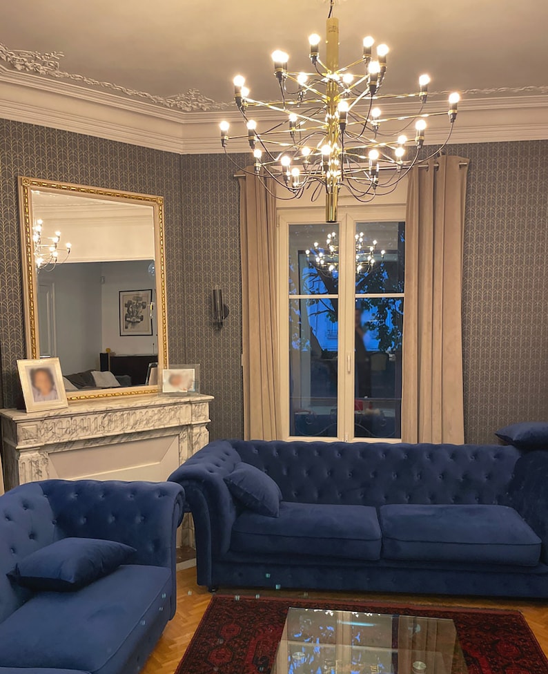 May include: A living room interior with a large gold chandelier, two blue velvet sofas, a glass coffee table, and a fireplace with a marble mantel.