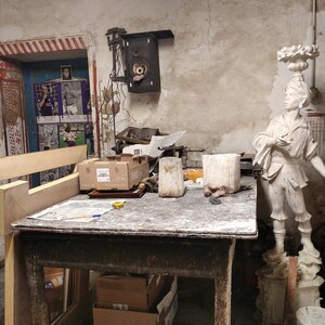 May include: A wooden work table with a white plaster statue of a person holding a scroll. There are two white ceramic containers and a cardboard box on the table. The table is in a room with a white wall and a speaker on the wall.