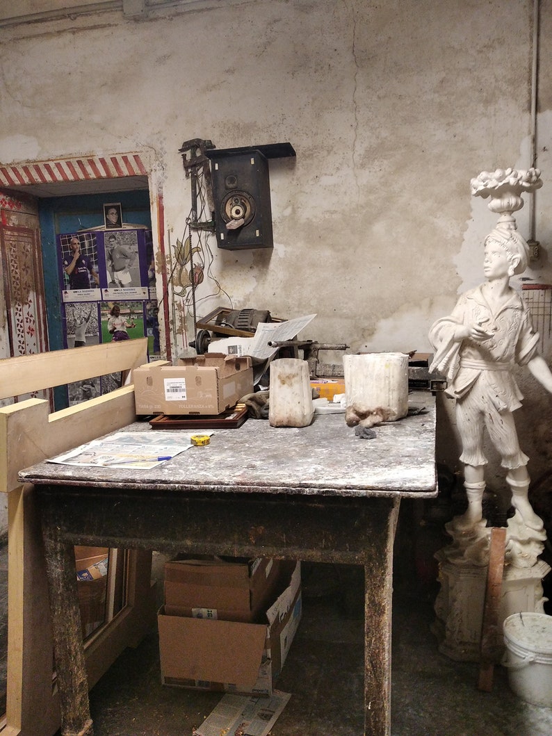 May include: A wooden work table with a white plaster statue of a person holding a scroll. There are two white ceramic containers and a cardboard box on the table. The table is in a room with a white wall and a speaker on the wall.