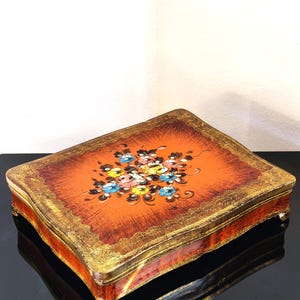 Op de afbeelding: Een rechthoekige houten doos met een feloranje bovenzijde voorzien van een handgeschilderd bloemenboeket in blauw, geel en roze. De randen en zijkanten zijn afgewerkt met een sierlijk, verweerd bladgoudpatroon en vier kleine, uitgesneden pootjes.