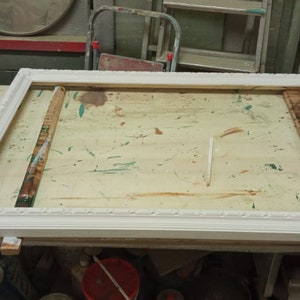 May include: A white wooden picture frame with a simple ornate design. The frame is sitting on a wooden table with paint splatters.