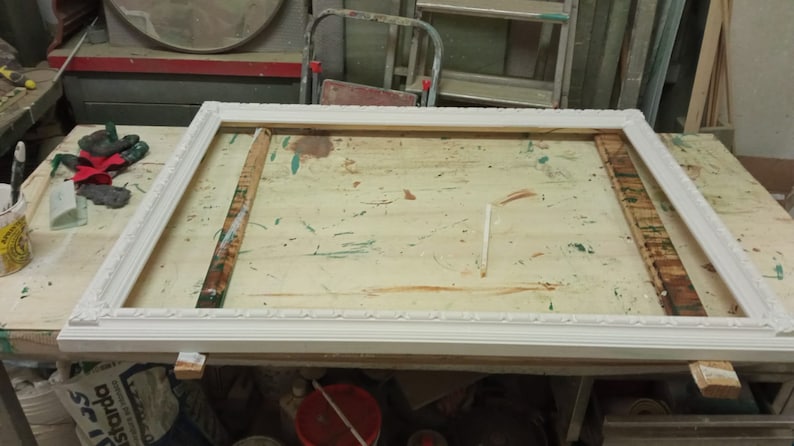 May include: A white wooden picture frame with a simple ornate design. The frame is sitting on a wooden table with paint splatters.