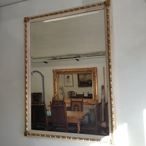 May include: A white wooden frame with gold accents surrounds a large mirror reflecting a room with furniture and other framed artwork.