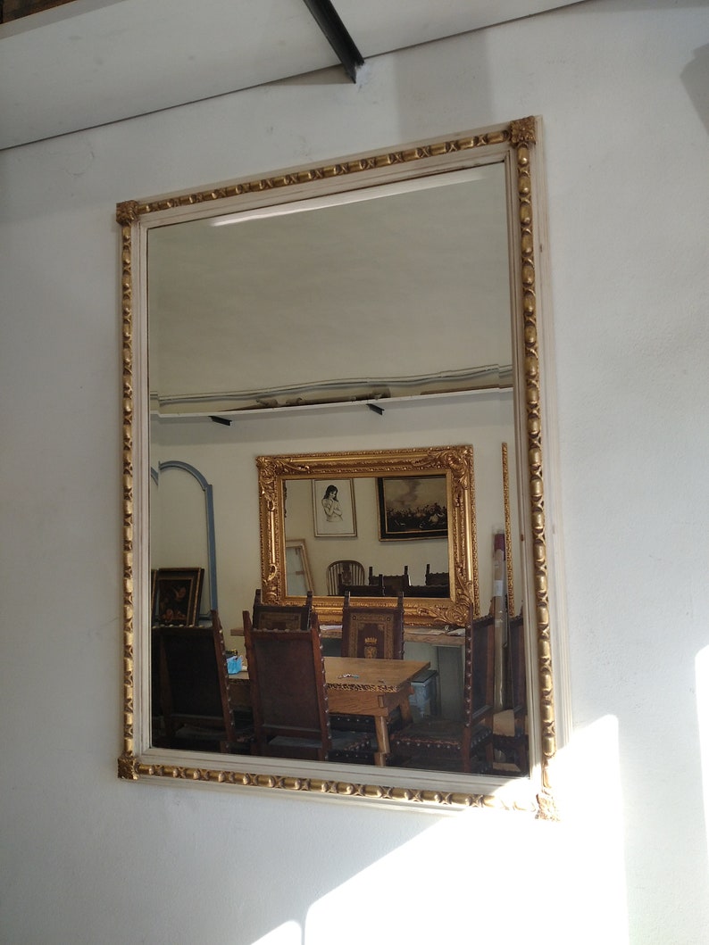 May include: A white wooden frame with gold accents surrounds a large mirror reflecting a room with furniture and other framed artwork.