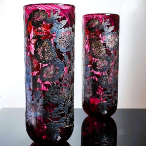 Puede incluir: Dos jarrones cilíndricos altos, de un profundo color magenta, con intrincados diseños florales en plata y gris. Los jarrones tienen un acabado brillante y se reflejan en una superficie oscura, creando un efecto simétrico. Los patrones florales añaden elegancia.