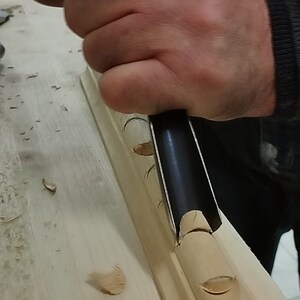 May include: A hand using a wood carving tool to create a decorative design on a piece of wood. The wood is light brown and the tool is black with a wooden handle.