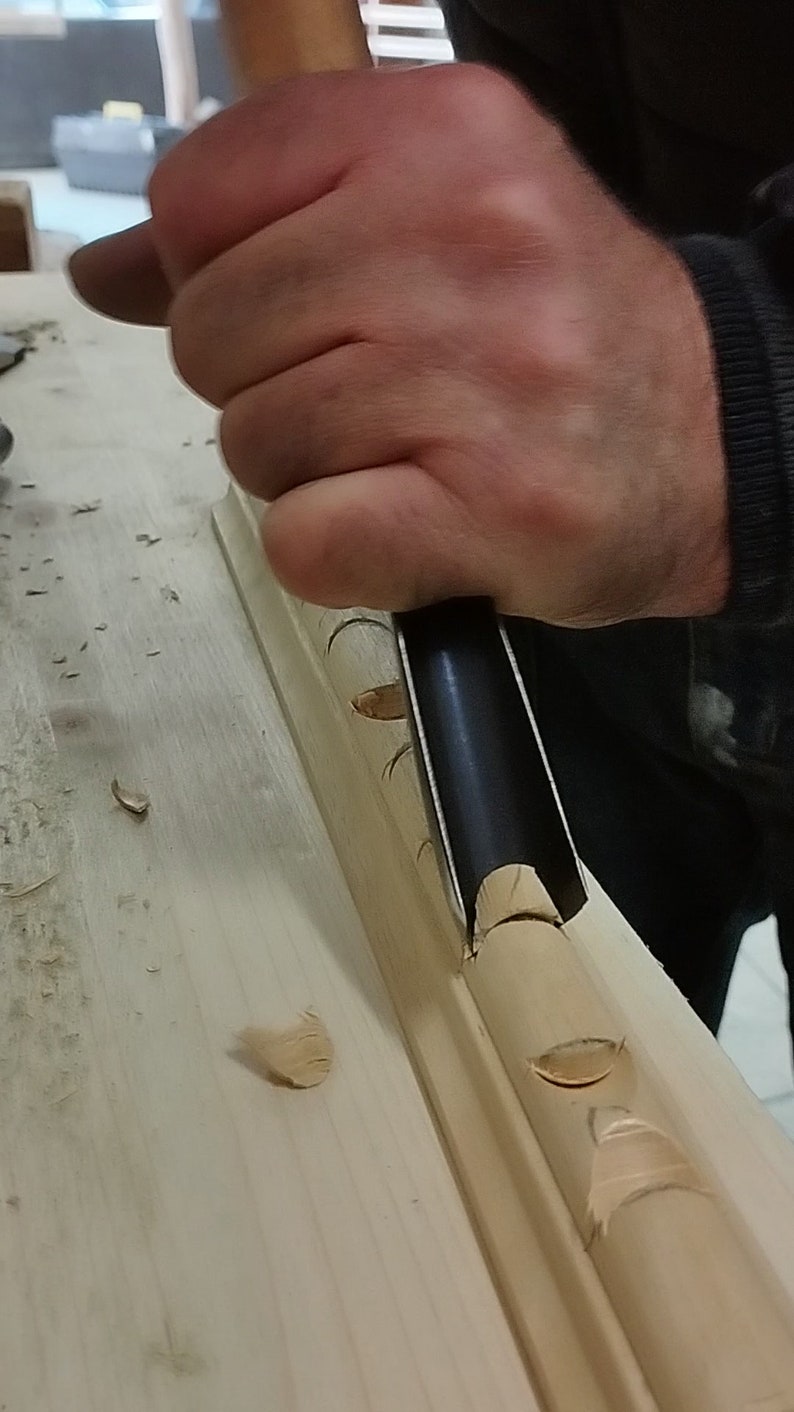 May include: A hand using a wood carving tool to create a decorative design on a piece of wood. The wood is light brown and the tool is black with a wooden handle.