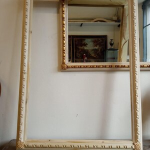 May include: Two ornate picture frames with a white wood finish and gold accents. The larger frame is empty, while the smaller frame has a mirror reflecting a workshop.