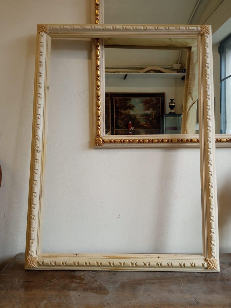May include: Two ornate picture frames with a white wood finish and gold accents. The larger frame is empty, while the smaller frame has a mirror reflecting a workshop.