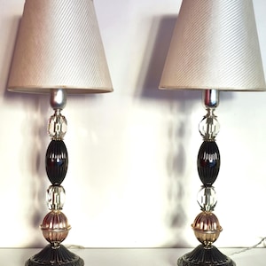 May include: A pair of table lamps with white fabric lampshades. The bases are black with silver accents and feature a beaded design with clear and gold glass beads.