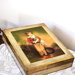 May include: A rectangular wooden box with a distressed gold finish featuring a printed illustration on the lid. The image depicts a child in a red jacket holding a small white dog, standing in a garden setting with a dark hat resting on the ground.