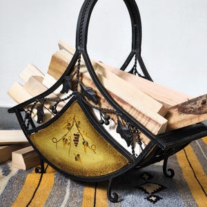 Op de afbeelding: Zwarte smeedijzeren houtmand met een gebogen handvat en decoratieve blad- en druivenaccenten. De mand heeft een geschilderd paneel met een wijnstokontwerp en is gevuld met brandhout.