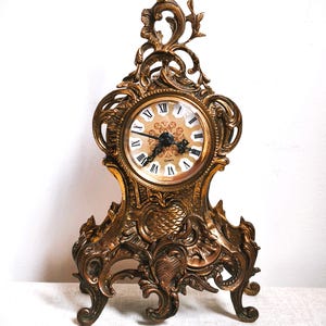 May include: Ornate gold-colored quartz clock with Roman numerals on the face. The clock features an elaborate, scrolling design and stands on four decorative feet. The clock face has a floral design in the center.