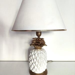 Puede incluir: Lámpara de mesa de cerámica blanca en forma de piña con una pantalla blanca con ribete dorado. La base de la lámpara tiene forma de piña con una parte superior dorada.