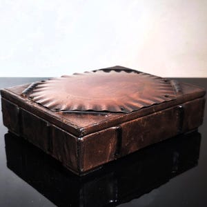 Puede incluir: Una caja rectangular de cuero marrón con una tapa decorativa en forma de sol. La tapa está hecha de un material marrón más claro y está remachada a la caja. La caja tiene una superficie texturizada.