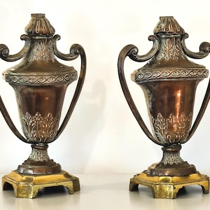 May include: A pair of antique bronze urns with ornate detailing. Each urn features a copper-colored body, silver accents, and curved handles. The base is a square shape with a gold finish. The urns are decorative.