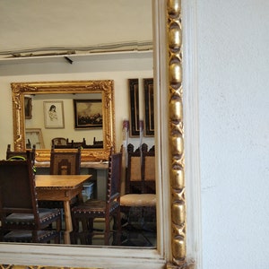 May include: A white mirror with a gold ornate frame. The mirror is reflecting a room with a wooden table and chairs.