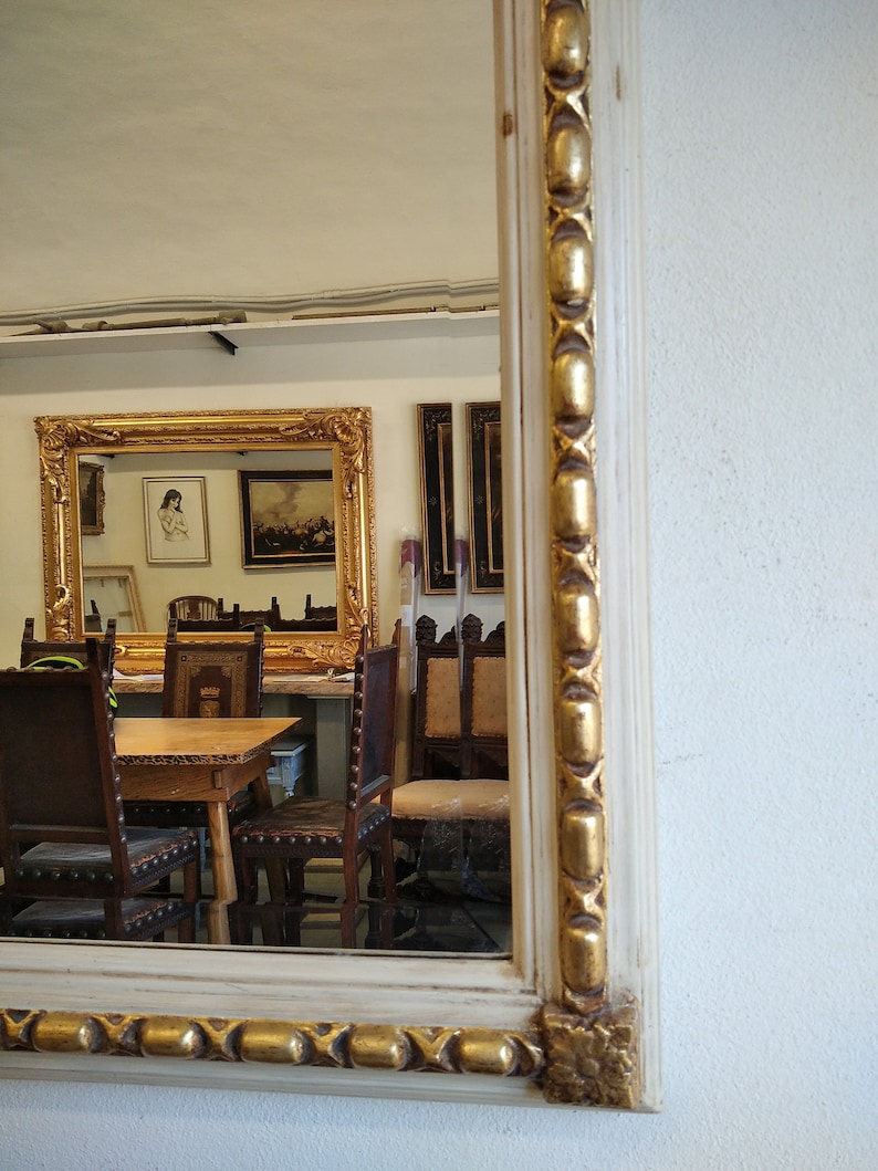 May include: A white mirror with a gold ornate frame. The mirror is reflecting a room with a wooden table and chairs.