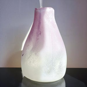 May include: A frosted glass vase with a rounded body and a flared top. The vase has a gradient of color, transitioning from a soft lavender at the top to a white base. The surface has a textured, speckled appearance, and it sits on a reflective surface.