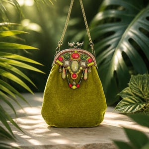 Pode incluir: Uma bolsa de mão em veludo verde-oliva com alça de corrente dourada e fecho de metal em forma de dois pássaros. A parte superior é decorada com contas coloridas, cabochões vermelhos e verdes e detalhes pendentes em tons de rosa e verde.