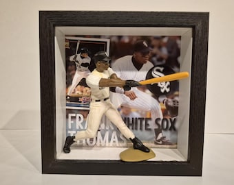 Pode incluir: Uma caixa de sombra emoldurada com uma figura de beisebol em posição de rebatida. A figura está vestindo um uniforme branco e preto. O fundo inclui um cartão de beisebol e uma foto de um jogador de beisebol. O texto "FRANK THOMAS" e "WHITE SOX" são visíveis.