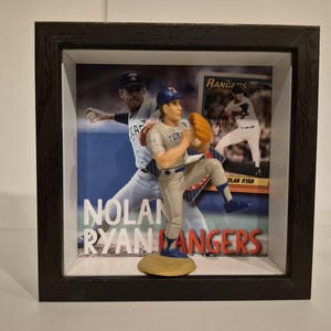 May include: A framed baseball display featuring a figurine of Nolan Ryan in a Texas Rangers uniform. The display includes a background image of Ryan pitching and the text "Nolan Ryan Rangers". The frame is black.