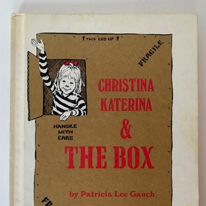 May include: A children's book cover featuring the title "Christina Katerina & The Box" in red lettering. An illustration shows a girl waving from a cardboard box. The cover also includes the author's and illustrator's names, and "Fragile" labels.
