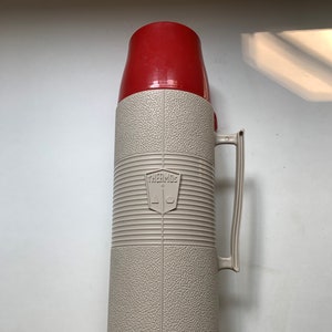 May include: A red and white thermos with a handle. The thermos has a textured surface and the brand name "THERMOS" is printed on the side.