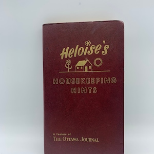 Peut inclure: Un livre bordeaux intitulé "Heloise's Housekeeping Hints" avec une illustration dorée d'une maison, d'un soleil et d'une fleur. Le livre est une publication du journal The Ottawa Journal.
