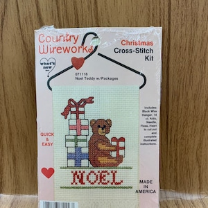 May include: A cross-stitch kit featuring a brown teddy bear sitting next to a stack of presents with the word "NOEL" stitched below. The kit includes floss, a needle, Aida cloth, a hanger, and instructions. Made in America.