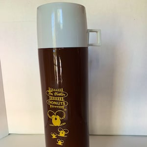 May include: A brown and white insulated thermos with a Tim Hortons Donuts logo and a cartoon donut character. The thermos has a white lid and a white handle.