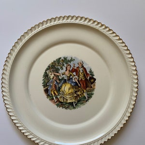 Puede incluir: Un plato de cerámica blanco con un borde dorado y una ilustración vintage de una pareja y un niño en un jardín.