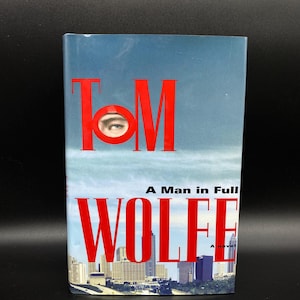 May include: A red book cover with the title "TOM WOLFE" and the words "A Man in Full" in white text. The cover features a cityscape with a close-up of a man's eye peeking out from behind the "O" in "TOM".