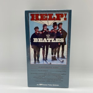 May include: A VHS tape cover for the movie "Help!" featuring the Beatles. The cover is blue and white with a picture of the Beatles in winter clothing. The text "Help!" is in red and white letters. The text "With The Beatles" is in white letters. The text "In Hi-Fi Digital Audio" is in black letters. The text "An MPI Home Video Release" is in black letters.