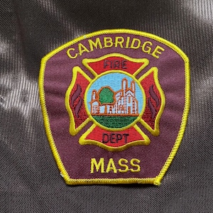 Op de afbeelding: Een geborduurde patch in de kleuren donkerrood en geel met de woorden "CAMBRIDGE FIRE DEPT MASS" en een rood kruis met een gebouw en vlammen.