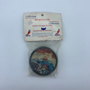 May include: A black and white hockey puck featuring a picture of Patrick Roy, a renowned hockey player, wearing a blue and white jersey with the NHLPA logo. The puck is contained within a plastic bag alongside a card that reads "Sport-Flash Collection Famous Hockey Players Les Grands Du Hockey".