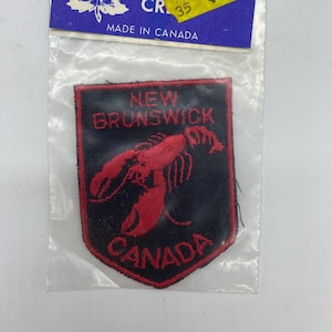 May include: A black and red embroidered patch featuring a red lobster and the words "NEW BRUNSWICK CANADA". The patch is in a clear plastic bag with a blue tag that says "CANADA" and "MADE IN CANADA".