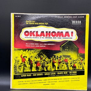 Peut inclure: Une pochette d'album de disque vinyle vintage pour la comédie musicale "Oklahoma!" avec une photo de la distribution de la production originale de New York. La pochette de l'album est jaune, rouge et noire avec le titre "Oklahoma!" en grandes lettres jaunes. La pochette de l'album comprend les noms des membres de la distribution, notamment Alfred Drake, Joan Roberts, Howard da Silva, Celeste Holm et Lee Dixon.