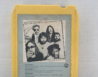 Può includere: Una vecchia cassetta a 8 tracce gialla dell'album "Minute by Minute" dei Doobie Brothers. La copertina dell'album presenta una foto in bianco e nero dei membri della band. La tracklist è stampata sotto l'immagine.