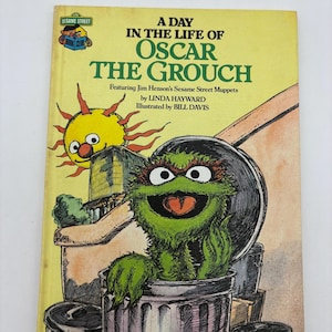 May include: A vintage children's book titled "A Day in the Life of Oscar the Grouch." The cover features a cartoon illustration of Oscar the Grouch in a trash can, with a smiling sun and the Sesame Street logo. The book is by Linda Hayward and illustrated by Bill Davis.