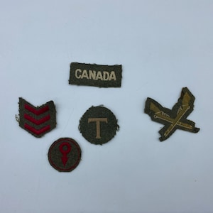 Może przedstawiać: Pięć wojskowych naszywek, w tym naszywka z napisem "CANADA" białymi literami, naszywka z trzema czerwonymi szelkami, naszywka z czerwoną szpilką, naszywka z białym "T" i naszywka z dwoma skrzyżowanymi złotymi karabinami.