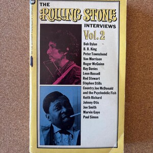 Könnte beinhalten: Ein Vintage-Taschenbuch mit dem Titel "The Rolling Stone Interviews Vol. 2". Das Cover zeigt Bilder von Bob Dylan und einem Mann, der eine Zigarette raucht. Das Cover des Buches ist gelb mit schwarzem und rotem Text.