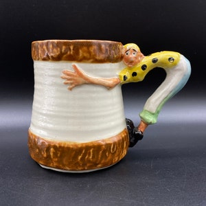 May include: A ceramic mug with a brown and white tree trunk design. The handle is a sculpted figure of a person with a yellow shirt, blue pants, and black shoes.