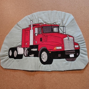 May include: Embroidered patch featuring a red semi-truck with black tires and a white exhaust stack. The truck is set against a light green background with a stitched border. The patch is likely for a jacket or bag.