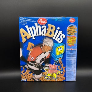 May include: A blue box of Alpha Bits cereal featuring a hockey player wearing a white and black jersey with the number 8 on the back. The box has a free collectible card offer with a Pinnacle logo. The text on the box reads "No Artificial Flavours" and "Sans Aromes Artificiels".